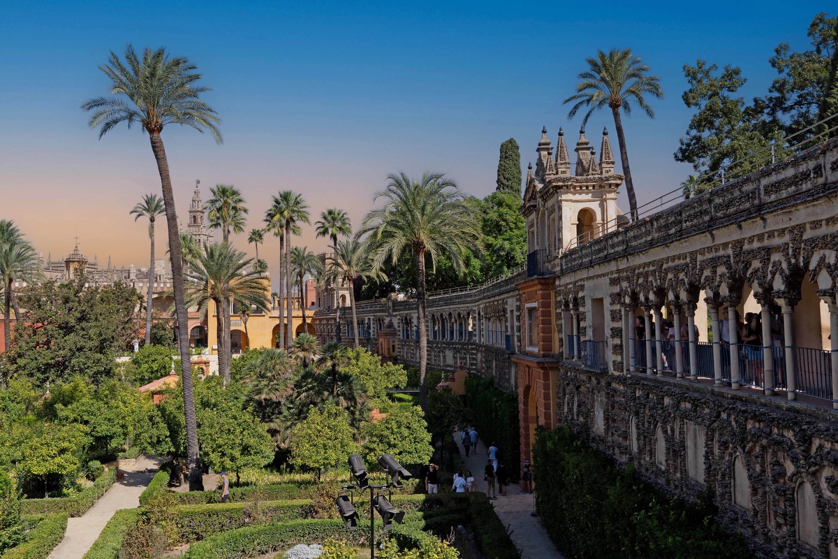 The gardens of the Real Alcázar in Seville at golden hour, with palm trees and the Giralda tower in the distance